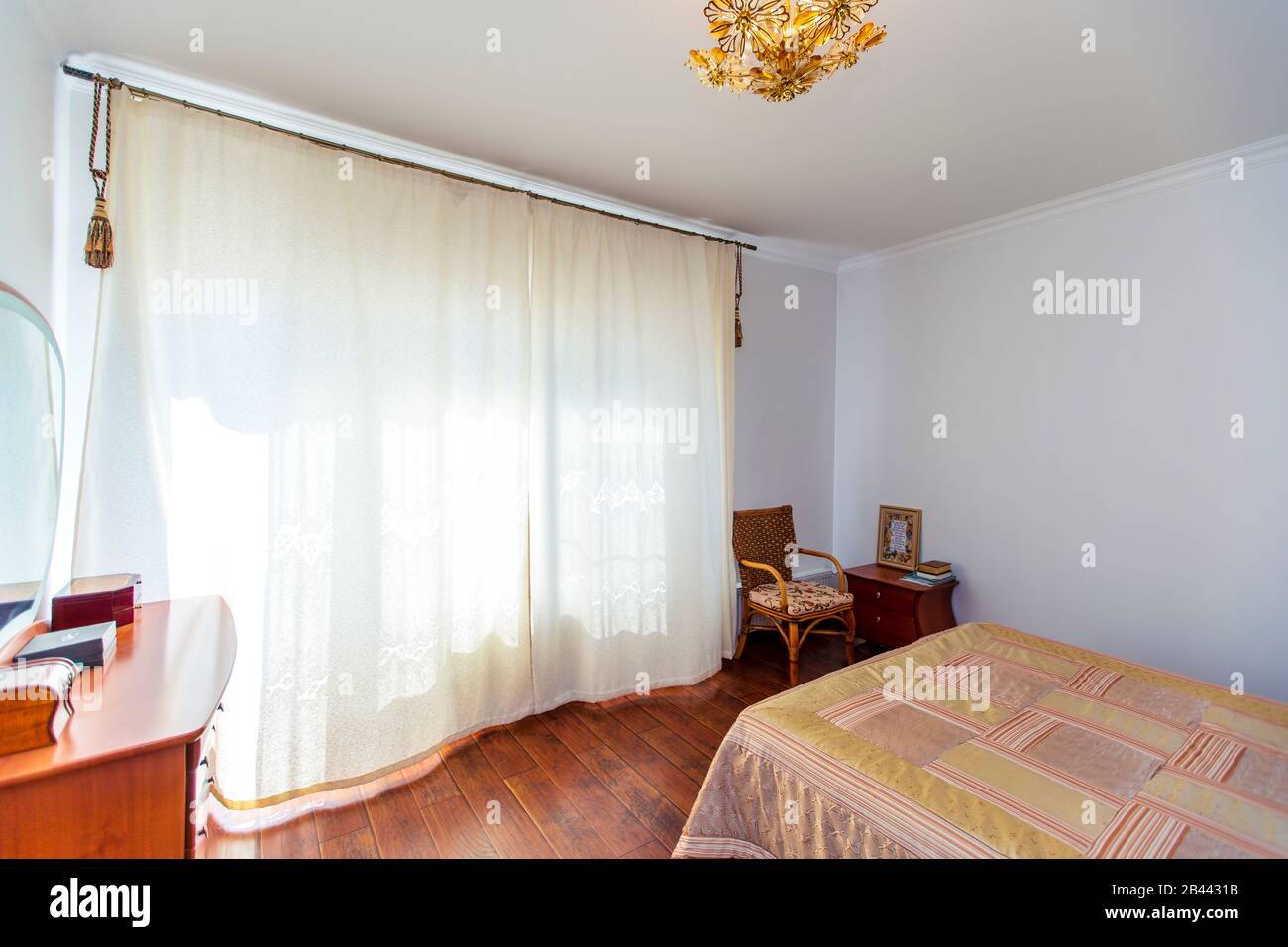 Ampia camera da letto con letto in mogano, sedie in vimini e elegante toeletta con specchio semicircolare. Il letto ha una coperta beige in piazze. Foto Stock