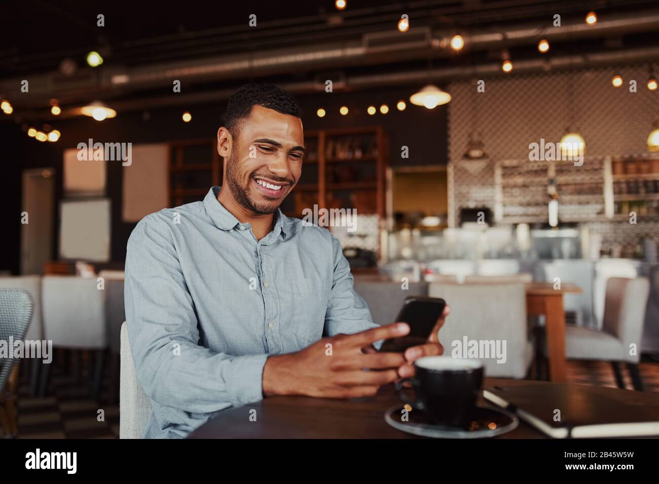 Allegro bel giovane africano che si rilassa in un moderno caffè usando il telefono cellulare mentre beve il caffè Foto Stock