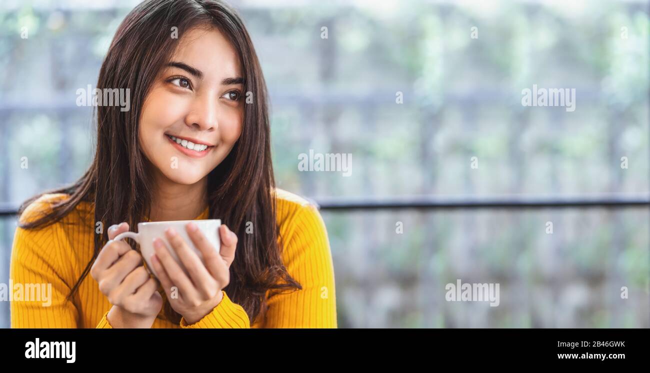 Asian giovane femmina che tiene una tazza di caffè nella moderna caffetteria o nello spazio di coworking accanto allo specchio della finestra, stile di vita e tempo libero con hobby e entrepr Foto Stock