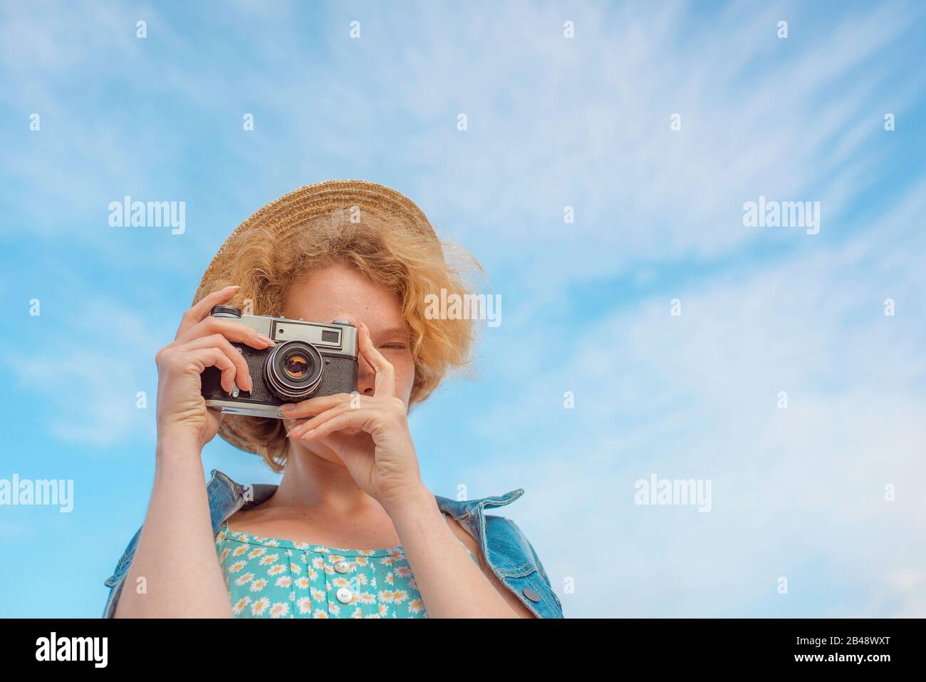 giovane donna curly redhead in cappello di paglia, sciess blu e giacca jeans in piedi con fotocamera vintage e scattare foto su sfondo blu cielo. Foto Stock