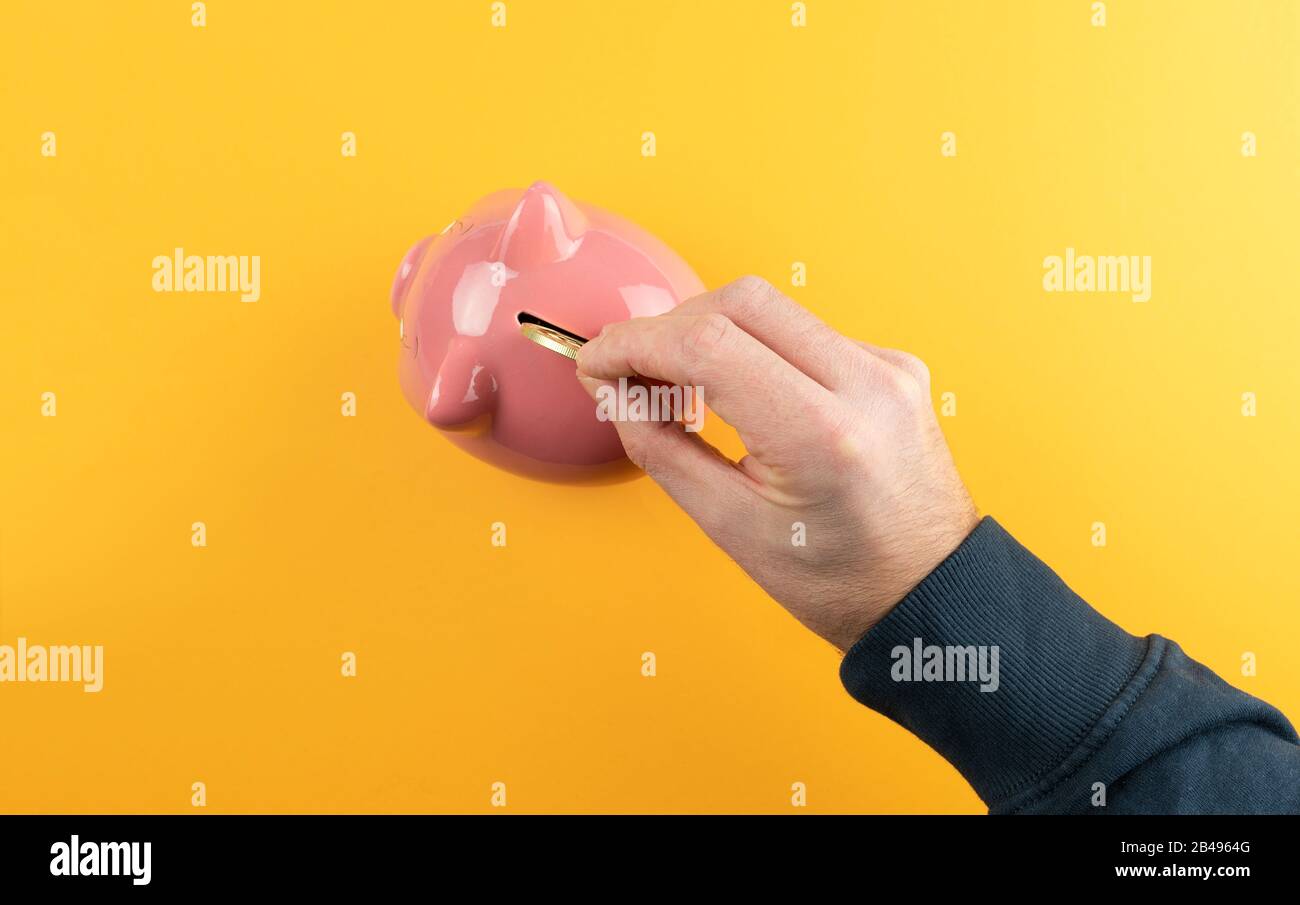 risparmio di denaro e il concetto di profitto, vista dall'alto della mano inserendo moneta dorata in banca rosa piggy contro sfondo arancione colorato Foto Stock