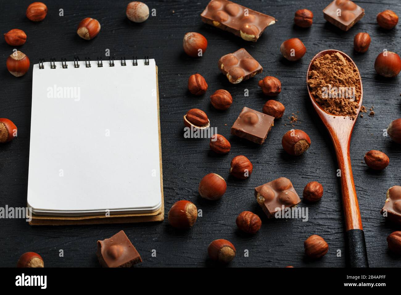 Cioccolato con nocciole, un taccuino con pagine bianche e un cucchiaio di legno con cacao su sfondo scuro, circondato da noci nel guscio e sbucciato. Foto Stock