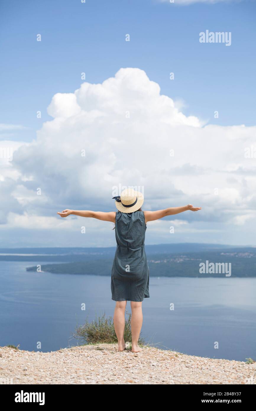 Donna in abito di lino e cappello è in piedi sulla costa della Croazia, diffonde le braccia e guarda al mare Foto Stock