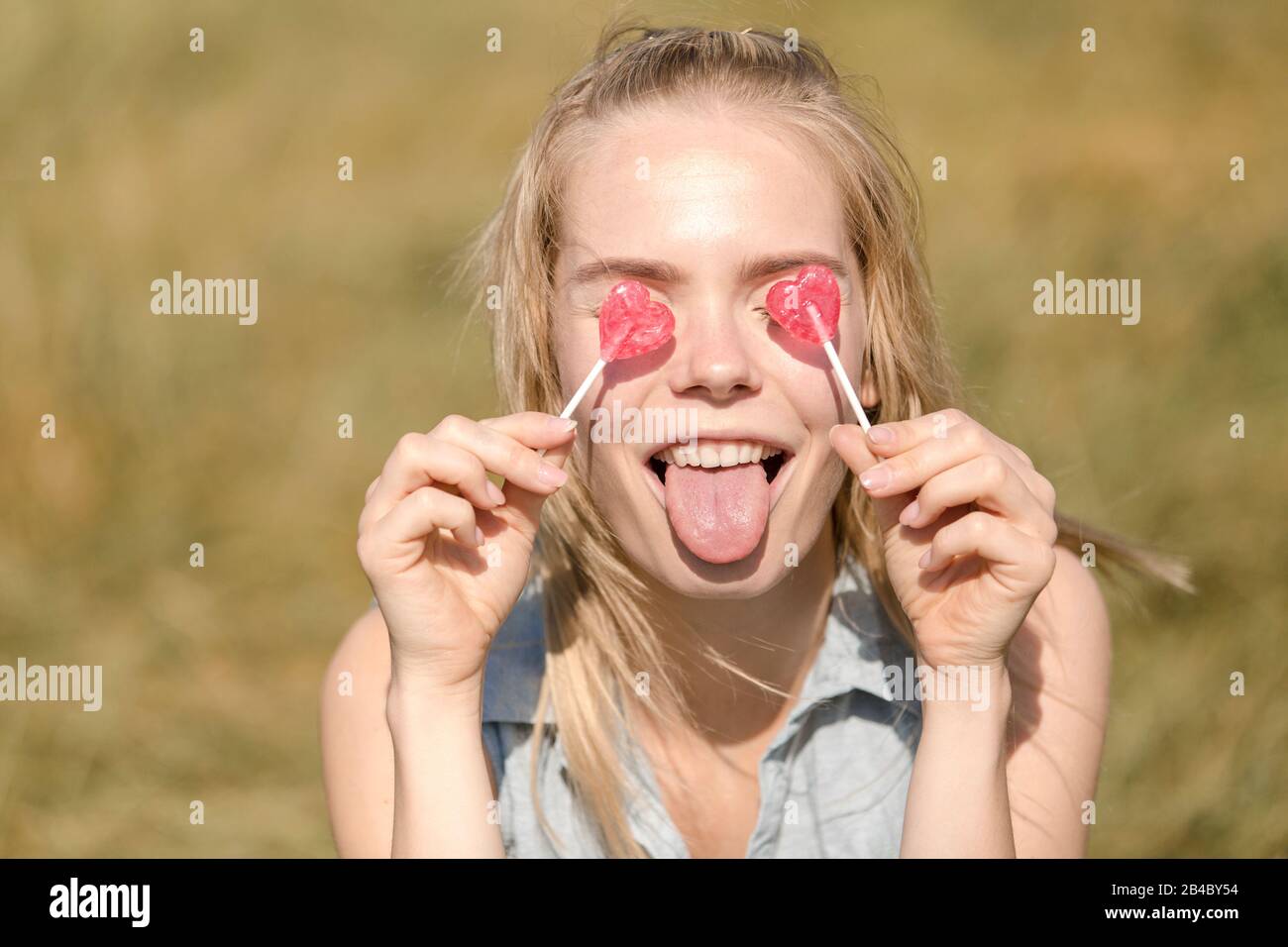 Junge Frau im Sommerkleid hält sich zwei Herzlutscher vor die Augen und streckt die Zunge raus Foto Stock