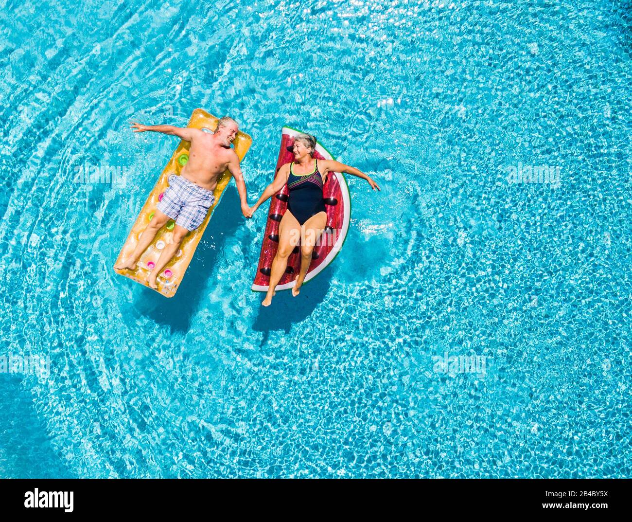 Sopra la vista verticale di persone anziani coppia prendere le mani con amore e divertirsi sulla piscina blu chiaro insieme godendo la vacanza estiva vacanza con il letto gonfiabile di colore alla moda lilos Foto Stock