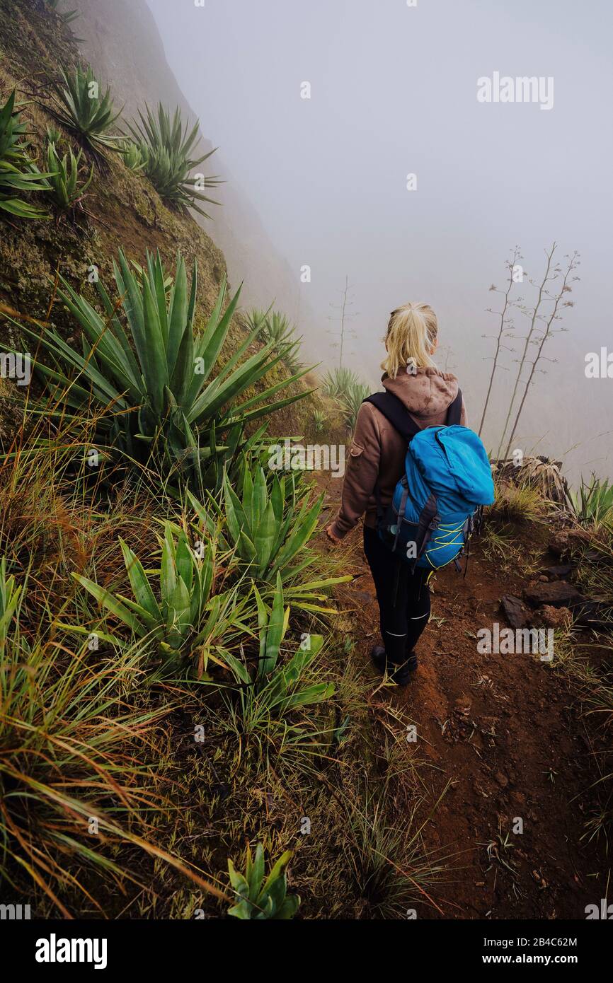 Viaggiatore femmina che rimane sul edhe del cratere inattivo del vulcano di insenatura sopra la valle verde nebbia sopraffondata con piante di yucca. Isola di Santo Antao a Capo Verde. Foto Stock