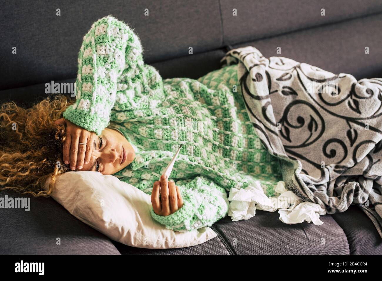 donna sola a casa sdraiata sul divano guardando il termometro con febbre alta temperatura - signora malata con capelli ricci utilizzando tessuti e coperta per la stagione invernale malattia fredda Foto Stock