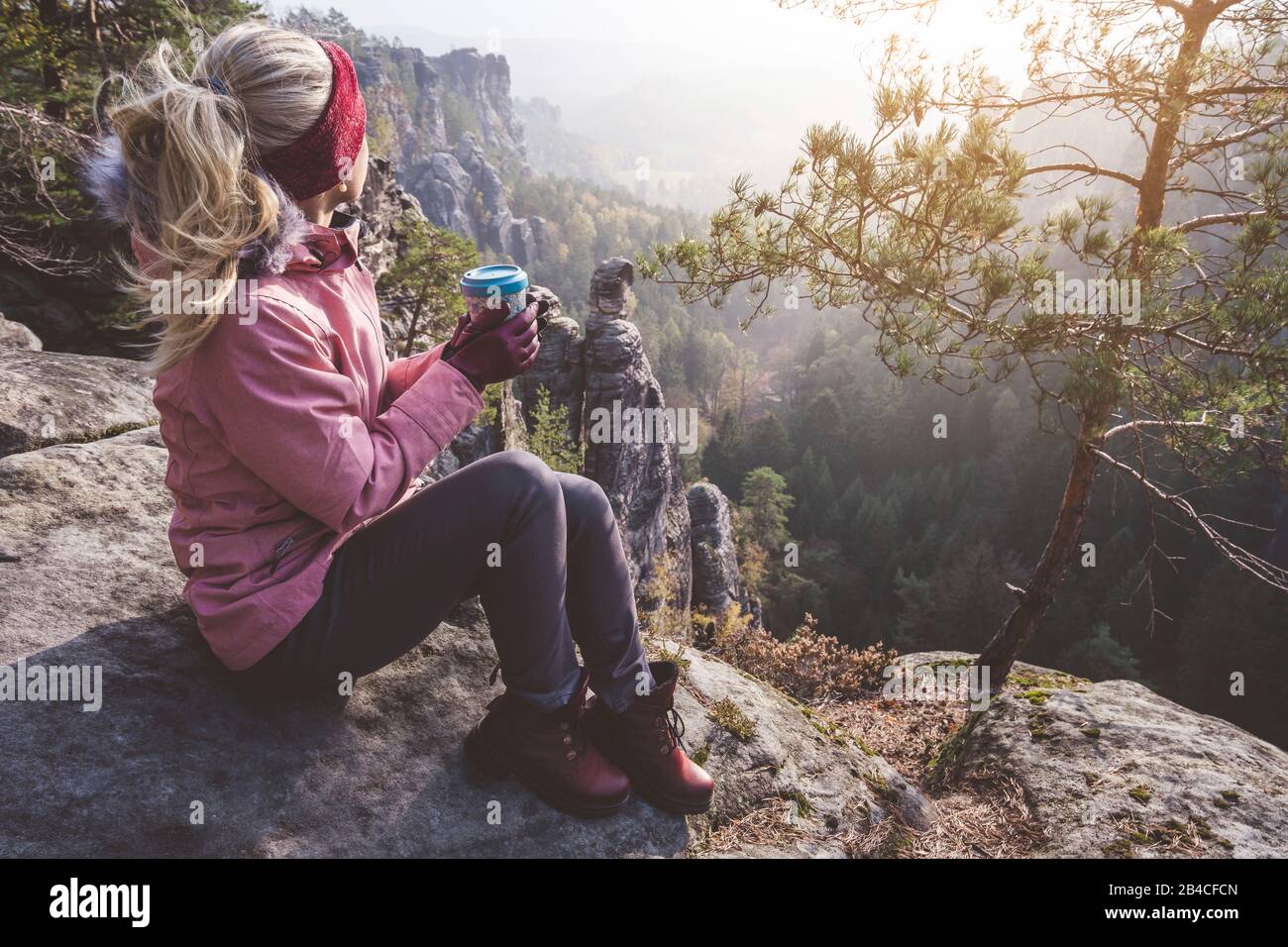 Biondo Frau mit Getränk in den Händen sitzt nach Wanderung auf einem Felsen und genießt die Aussicht, Reise-Lifestyle-Abenteuer-Konzept Foto Stock