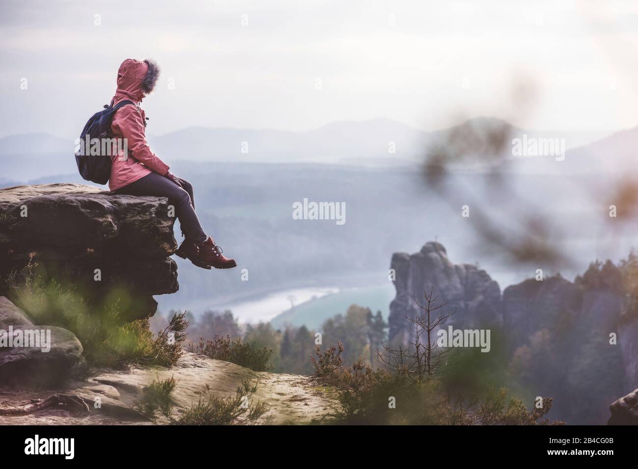 Junge Frau in winterlicher Kleidung mit Rucksack, sitzt am Rand der Klippe und genießt die Aussicht über Berge, tal und Fluss, Reise-Lifestyle-Konzept Foto Stock