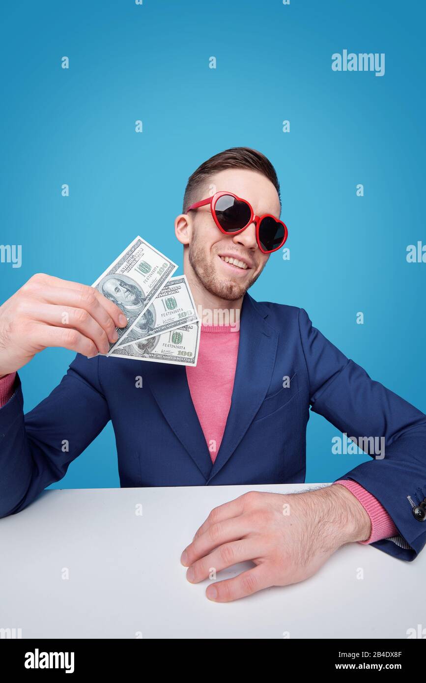 Giovane e di classe, ricco di giacca blu e occhiali da sole glamour con tre banconote in dollari mentre si siede alla scrivania Foto Stock