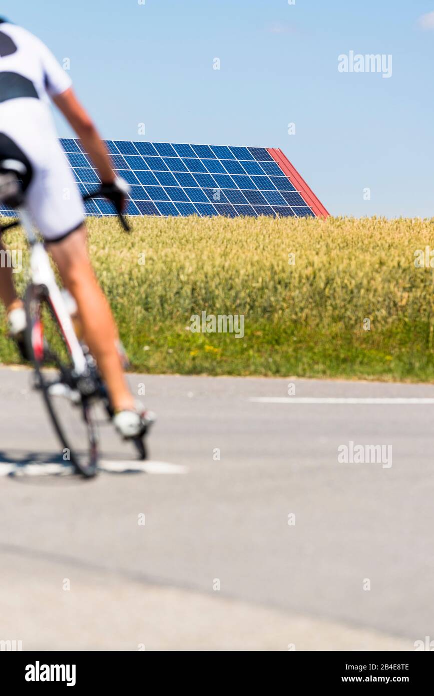 Rennradfahrer auf Landstrasse, Solaranlagen im Kornfeld unter blauem Himmel Foto Stock