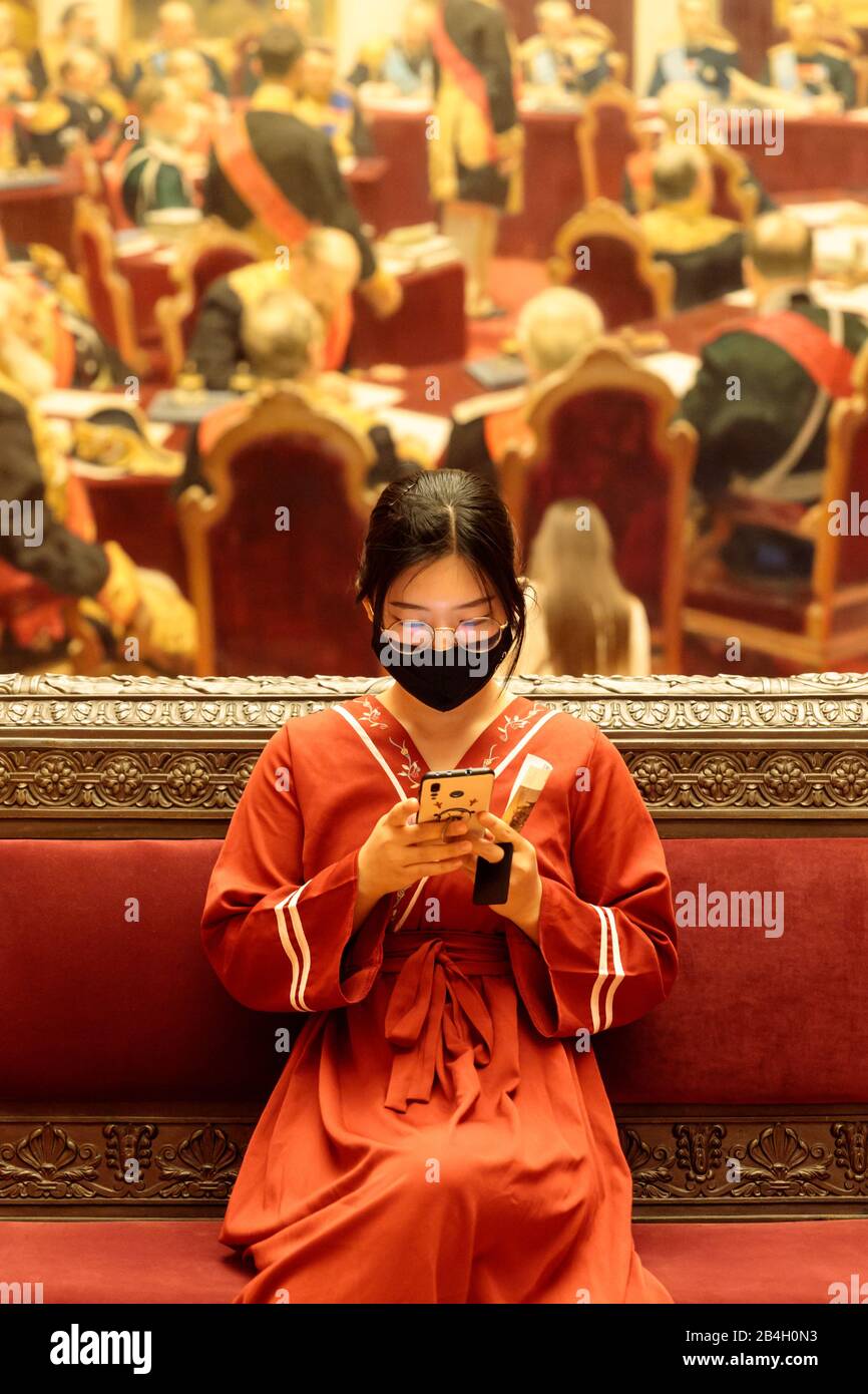 San Pietroburgo, Russia - Gen.2020: Giovane donna cinese in abiti nazionali indossando maschera chirurgica seduta nel museo, utilizzando lo smartphone. Nuovo tipo coronav Foto Stock