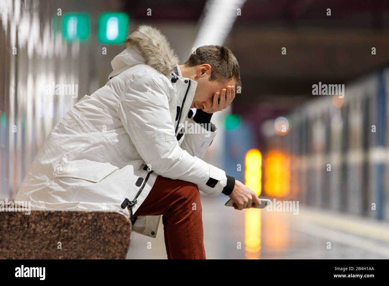 Piangendo l'uomo in parka bianco molto turbato, tenendo lo smartphone, ottiene cattive notizie, copre il suo viso con la mano, seduto su panchina nella stazione della metropolitana. Problema in Foto Stock