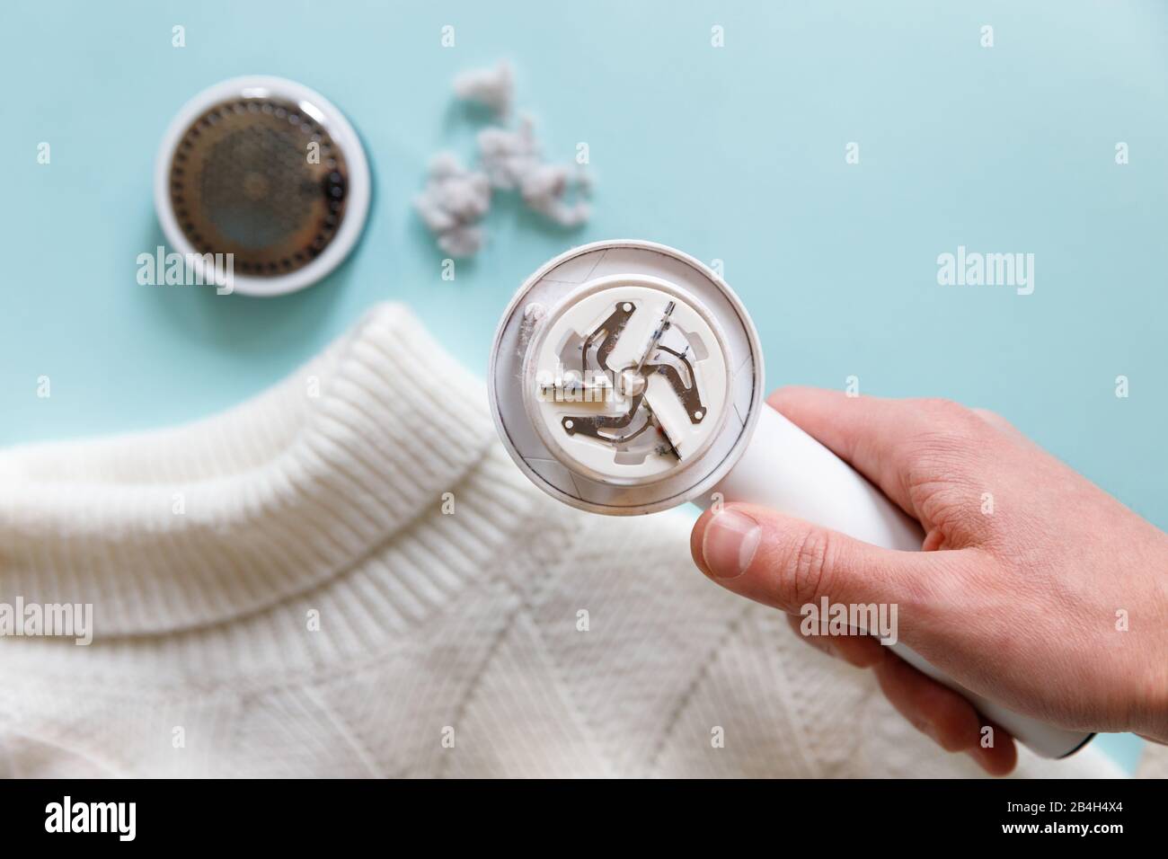 Mano dell'uomo che tiene un smacchiatore di pelucchi da un maglione bianco di lana o acrilico. Dispositivo elettrico dopo la pulizia e raccolta lanugine/lanugine, backgrou in legno chiaro Foto Stock