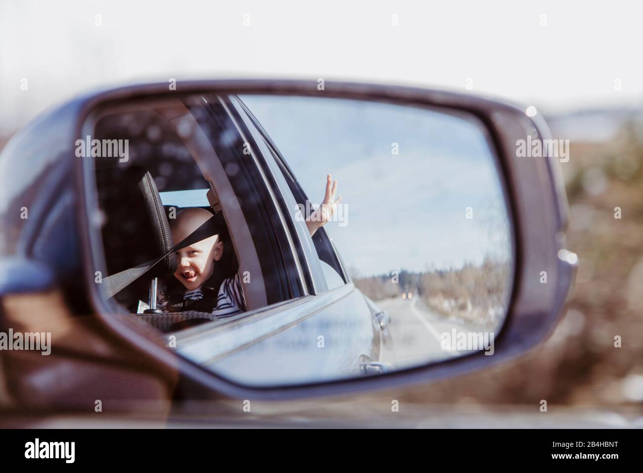 Specchio per auto girato di ragazzo in sedile posteriore con finestra a mano fuori il giorno di sole Foto Stock
