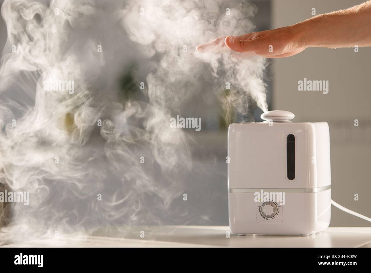 L'uomo tiene la mano sopra il diffusore di olio aromatico di vapore sul tavolo a casa, vapore dall'umidificatore dell'aria, spazio libero. Tecnologia a ultrasuoni, aumento di aria h Foto Stock