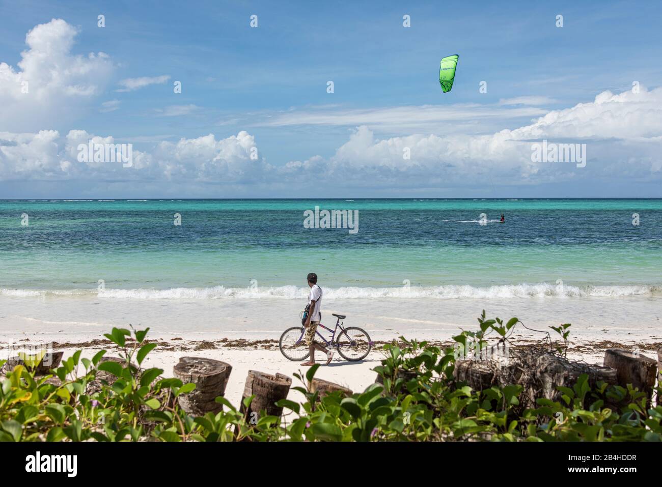 Zanzibar, Tanzania: Spiaggia da sogno sulla costa orientale di questa isola africana nell'Oceano Indiano. Islander spinge la sua bicicletta sulla spiaggia, kitesurfers in background. Foto Stock