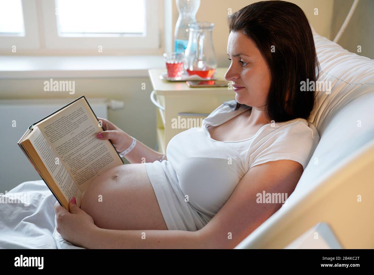Gravidanza ad alto rischio, donna incinta con libro a letto in ospedale, Foto Stock