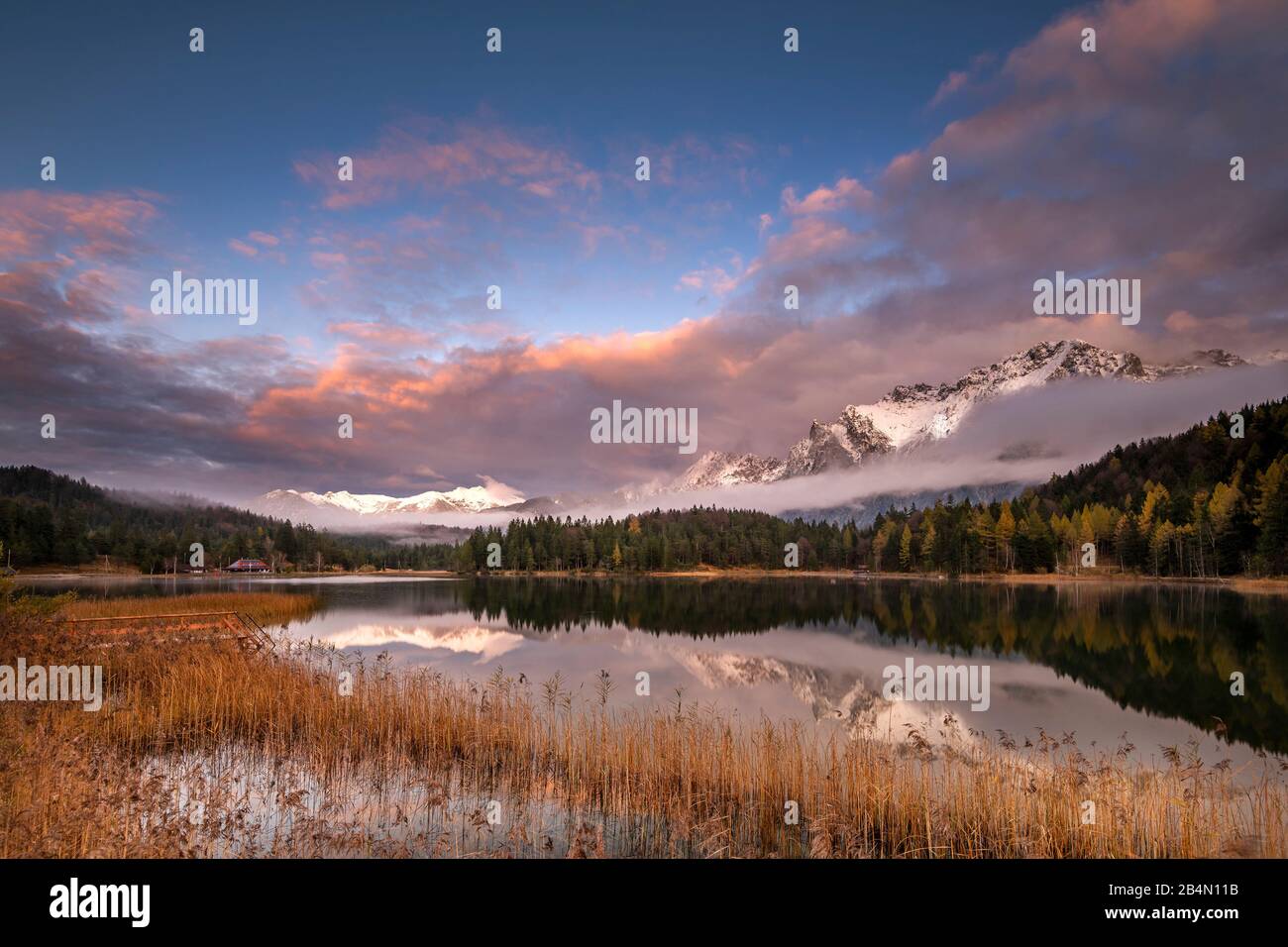 Abendrot am Ufer des Lautersee mit Schilf und Holzsteg. Im Hintergrund das westliche Karwendel mit Schnee, Nebel und Wolken. Foto Stock