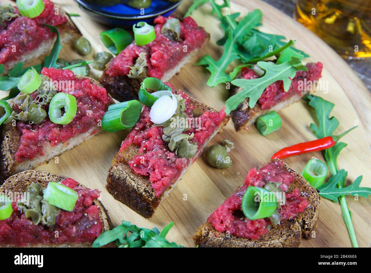 Sostanzioso spuntino tedesco: Piatto di legno isolato con carne di manzo arrosto tatar pane aglio, cipollotti, capperi e rucola insalata foglie Foto Stock
