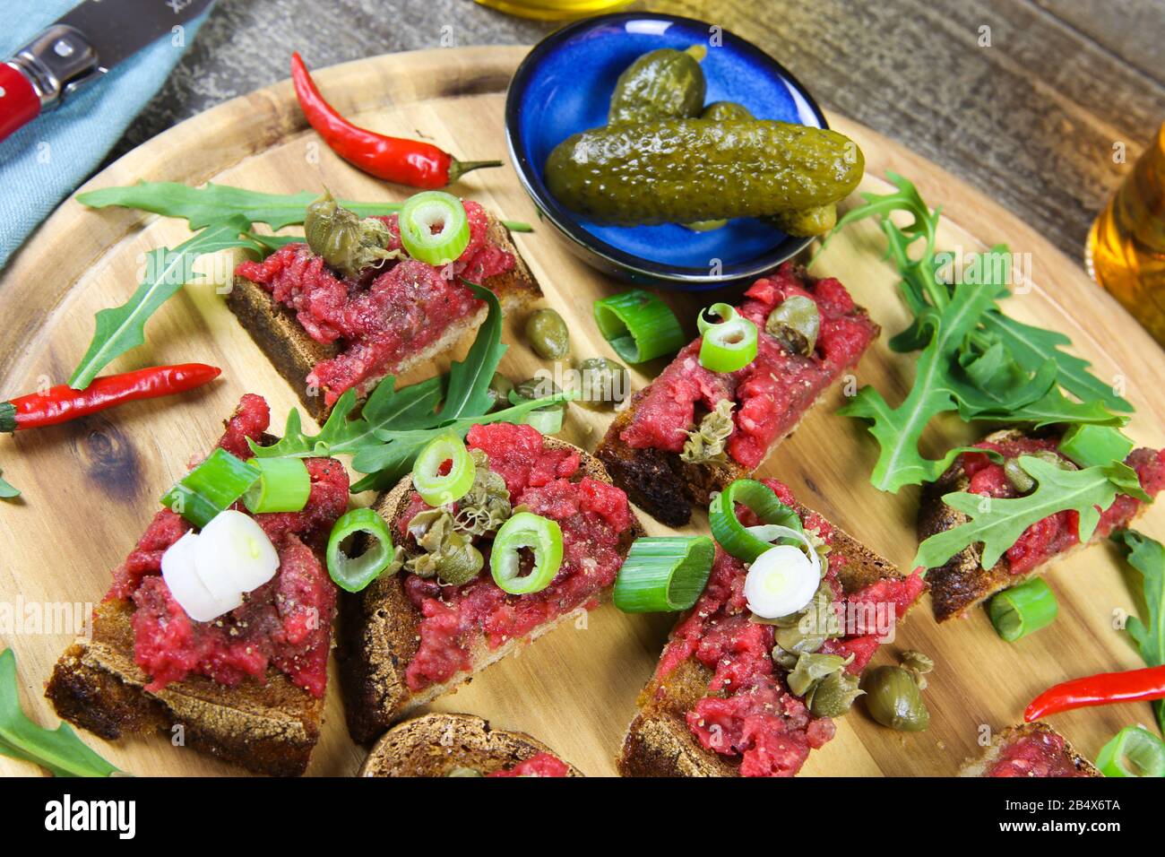 Sostanzioso spuntino tedesco: Piatto di legno isolato con carne di manzo arrosto tatar pane aglio, cipollotti, capperi, cetrioli sottaceto (cetriolini) e rucola sa Foto Stock