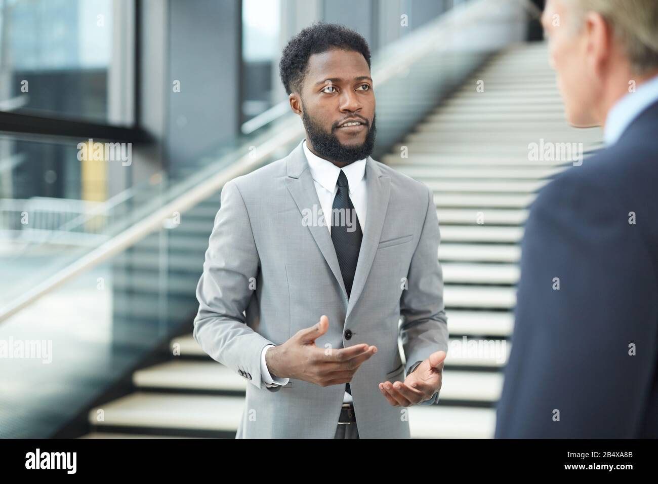 L'uomo nero elegante indossa un abito grigio davanti al suo collega irriconoscibile spiegandogli qualcosa, ritratto orizzontale Foto Stock