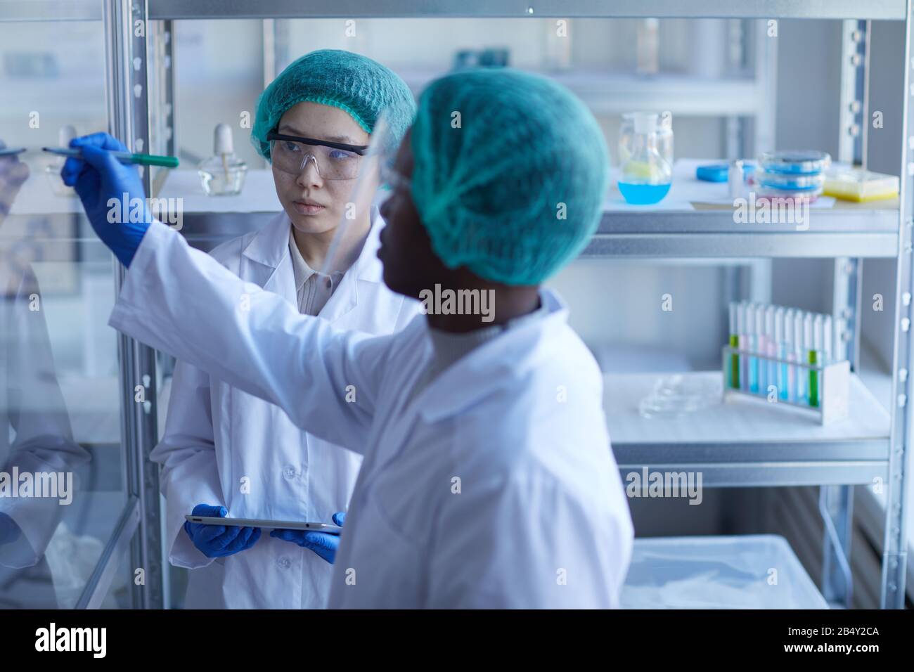 Ritratto orizzontale del lavoratore di laboratorio maschio nero formula di scrittura su parete di vetro, la sua collega asiatica femminile che lo guarda Foto Stock