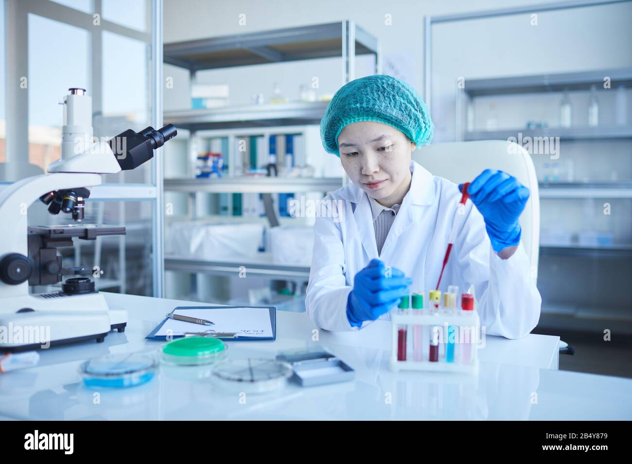 Ritratto orizzontale di giovane donna concentrata che indossa camice da laboratorio, guanti di protezione e cappello che esegue test medici, copia spazio Foto Stock