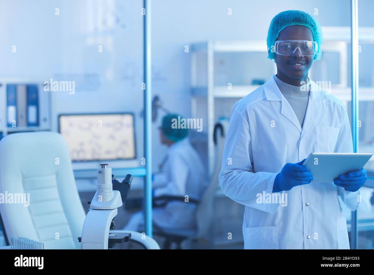 Ritratto orizzontale medio di bel giovane scienziato maschile indossando camice da laboratorio bianco, cappuccio medico e occhiali protettivi tenendo il tablet PC guardando ca Foto Stock