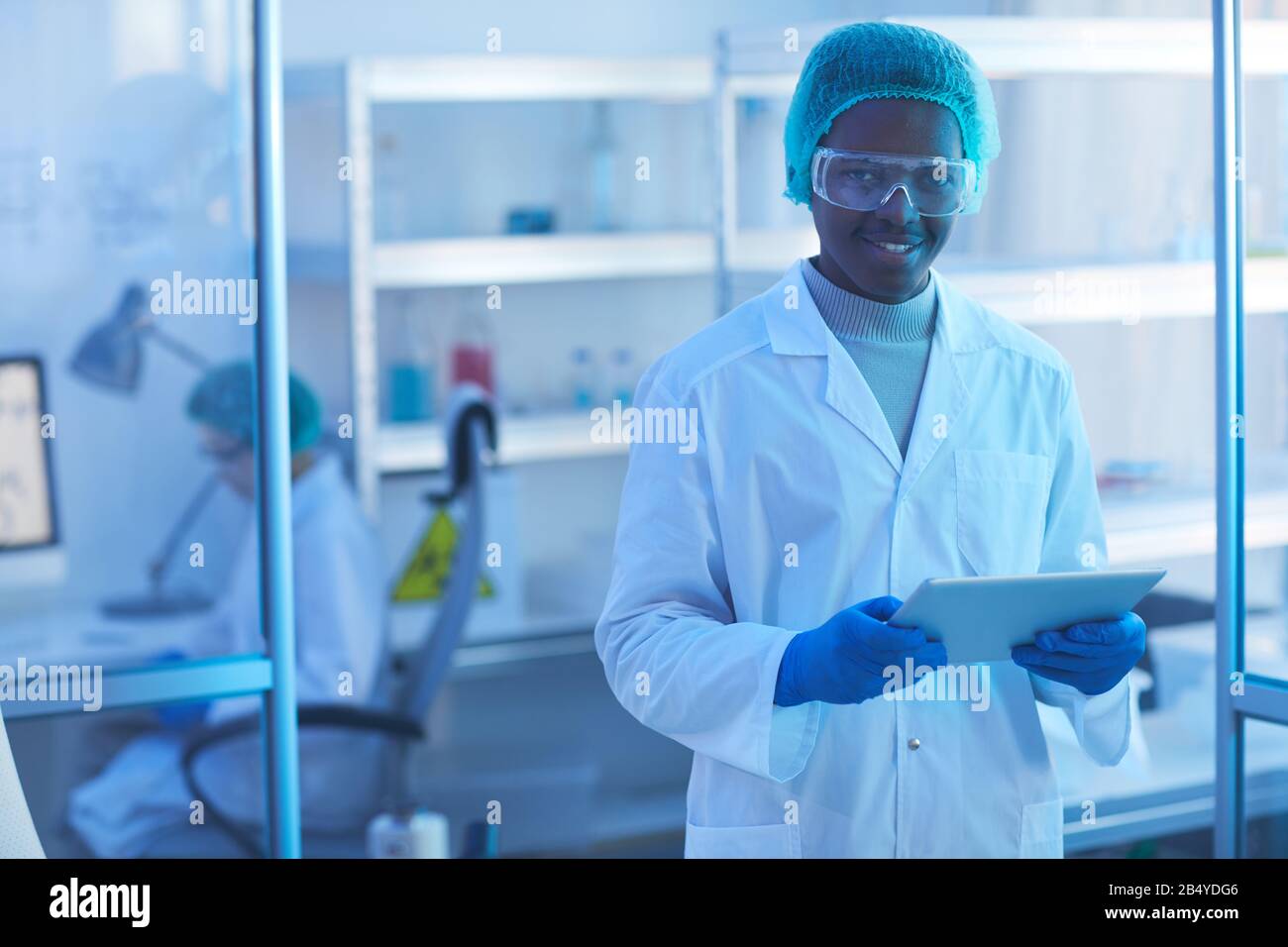 Ritratto orizzontale medio di giovane uomo allegro scienziato indossando camice da laboratorio bianco, cappuccio medico e occhiali protettivi tenere tablet PC guardando ca Foto Stock