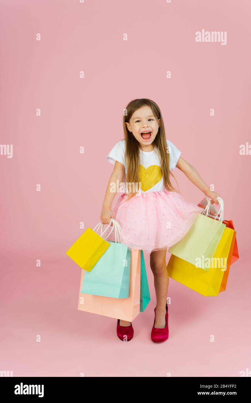 Bella ragazza carina piccola con capelli biondi lunghi, gonna tulle in piedi in madri grandi scarpe con pacchetti colorati isolato su sfondo rosa. Esprimere vere emozioni positive alla fotocamera. Foto Stock