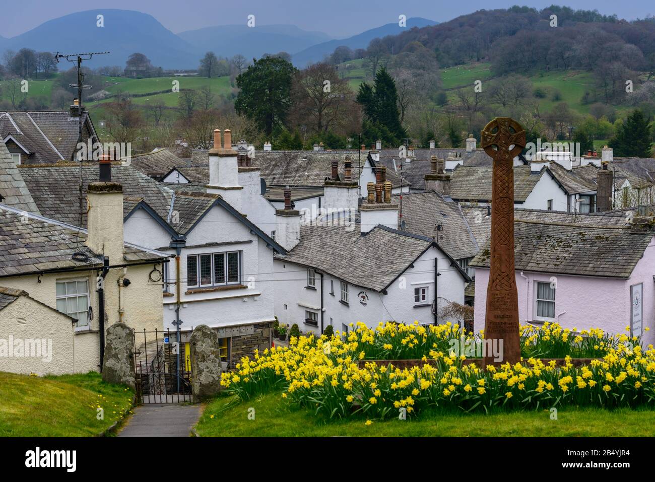 Un memoriale di guerra circondato da narcisi nei terreni di San Michele e Tutti gli Angeli Chiesa con il villaggio di Hawkshead, Cumbria, sullo sfondo. Foto Stock