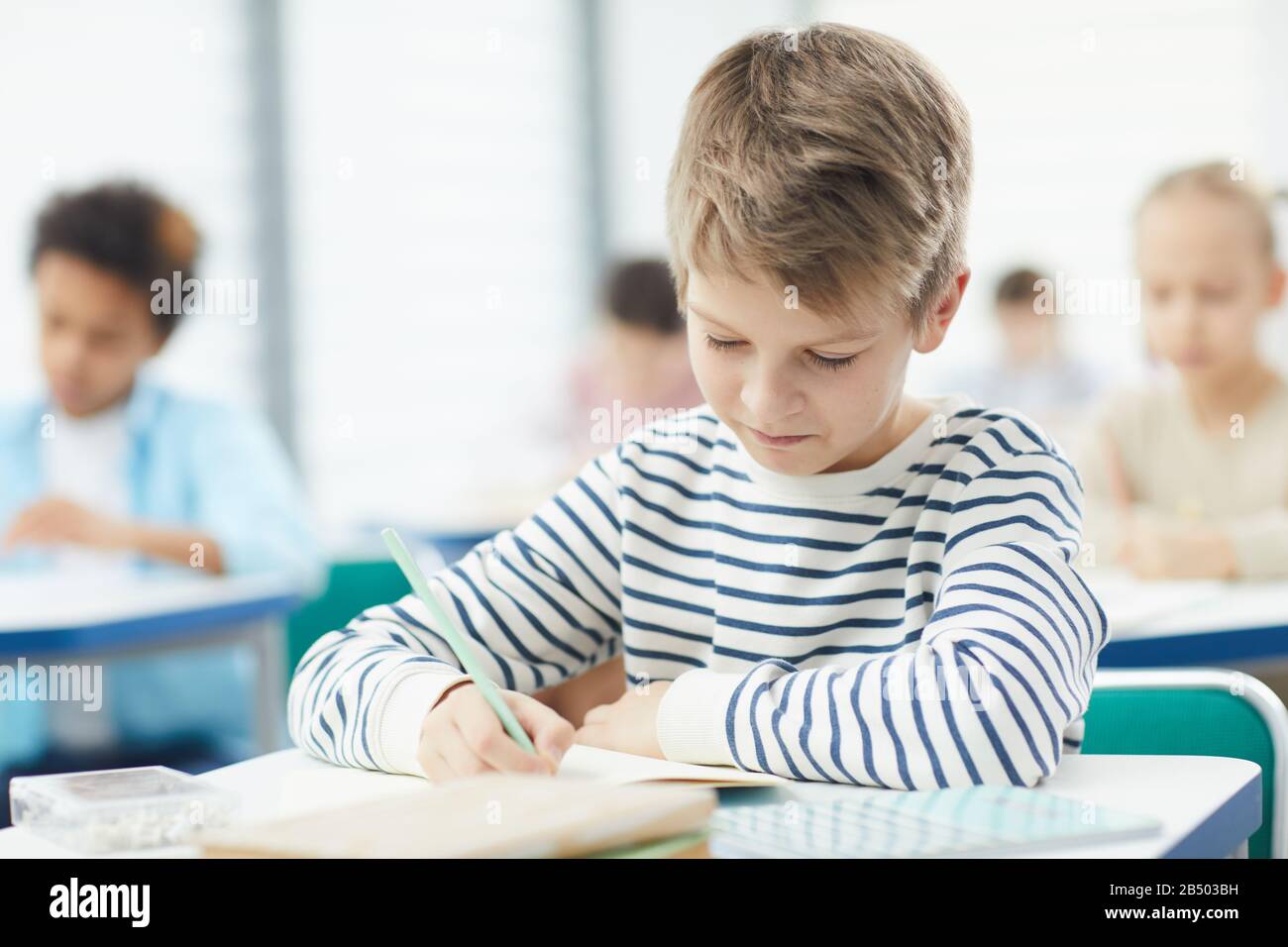 Ragazzo caucasico che indossa felpa a righe siatng alla scrivania in classe fare attività di lezione nel suo notebook, orizzontale medio primo piano ritratto, copia spac Foto Stock