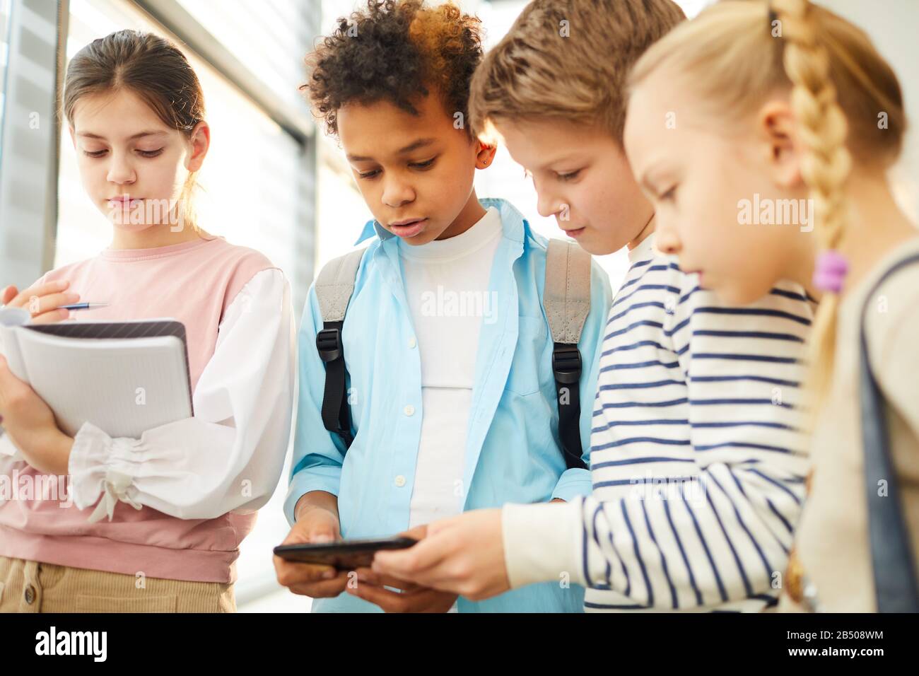 Due ragazzi e due ragazze che stanno insieme a guardare qualcosa in Internet sullo smartphone, il loro compagno di classe accanto a loro e leggere qualcosa in notebo Foto Stock