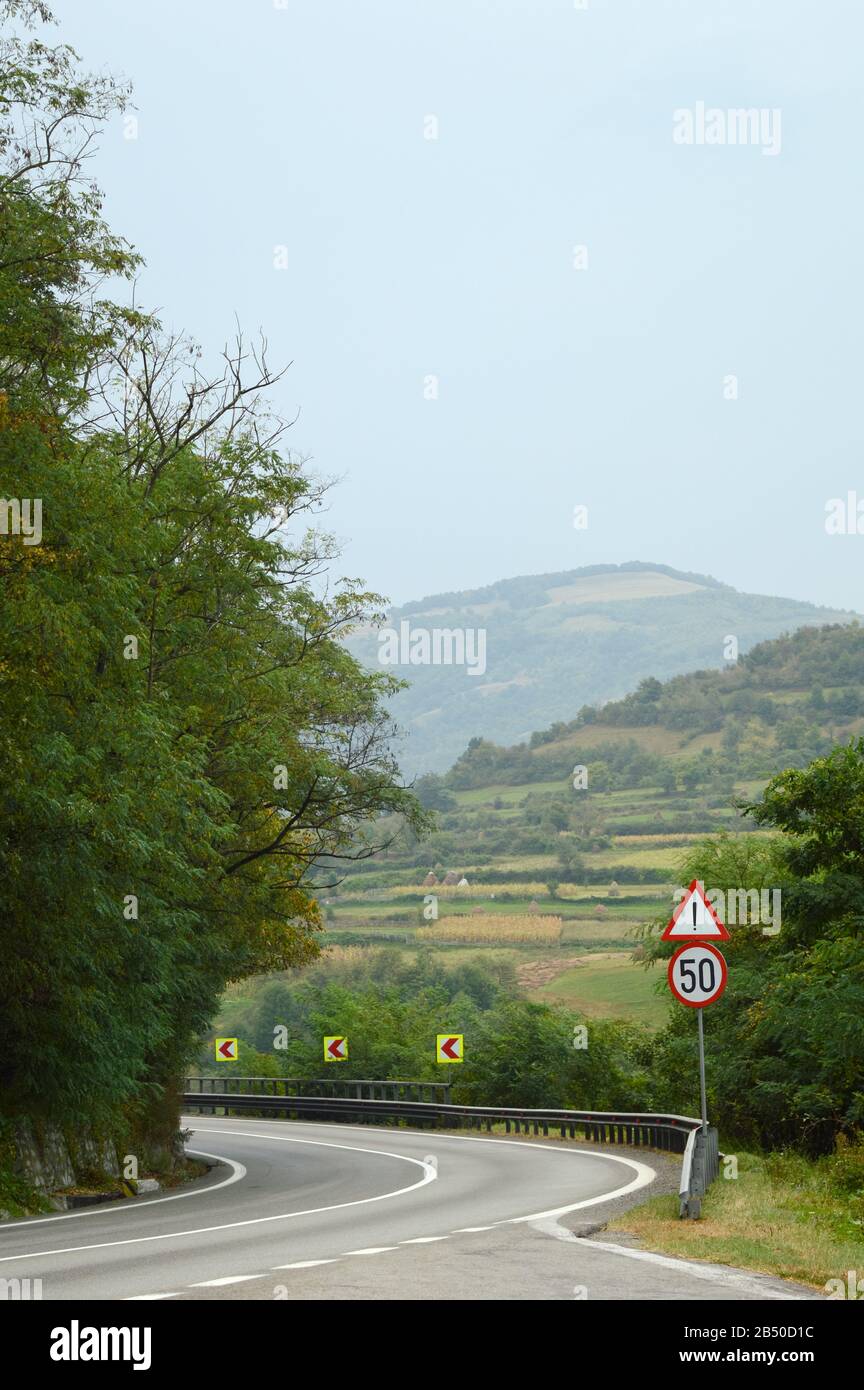 segnale stradale esclamativo e limite di velocità di 50 su una svolta pericolosa e sullo sfondo di un paesaggio rurale estivo con montagne Foto Stock