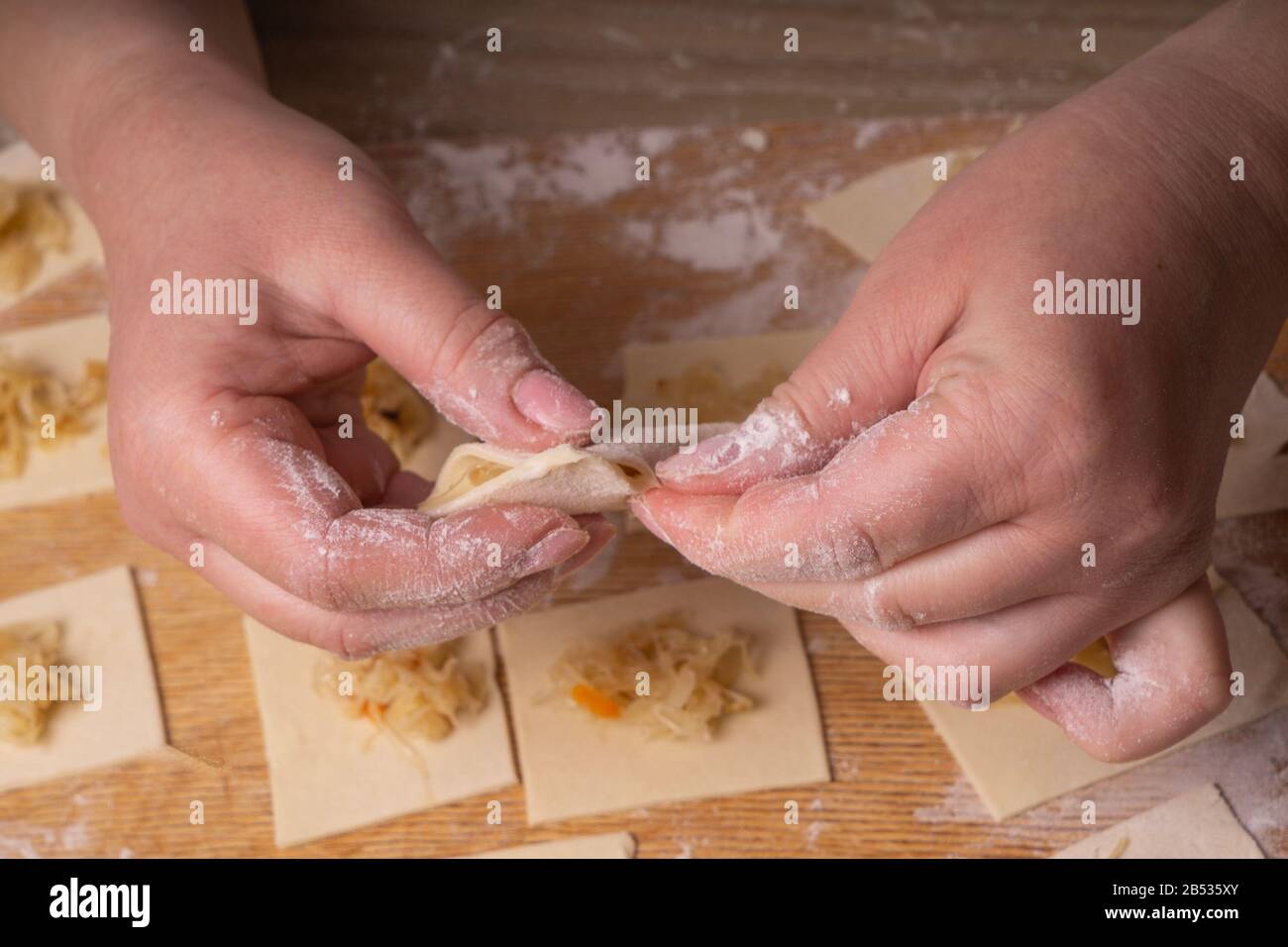 Una donna scolpisce gnocchi e ravioli da quadrati di pasta e cavolo. Pannello di taglio compensato, setaccio di farina di legno e perno di laminazione di legno - attrezzi per Foto Stock