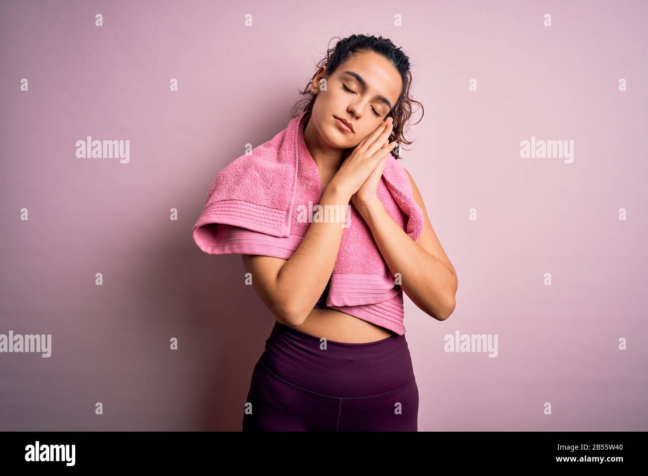 Giovane bella sportswoman con capelli ricci che fa sport utilizzando asciugamano su sfondo rosa dormire stanco sognare e posare con le mani insieme mentre Foto Stock