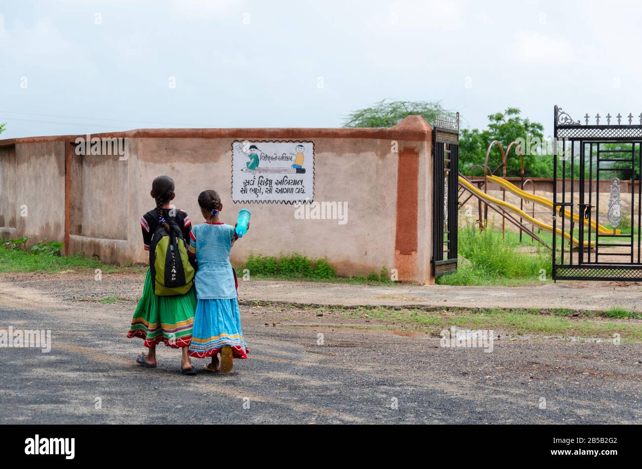 Due bambine che camminano con la borsa di scuola, entrando nel cancello della scuola contrassegnato da Sarva Shiksha Abhiyan logo a Gandhi Nu Gam, Kutch, Gujarat, India Foto Stock