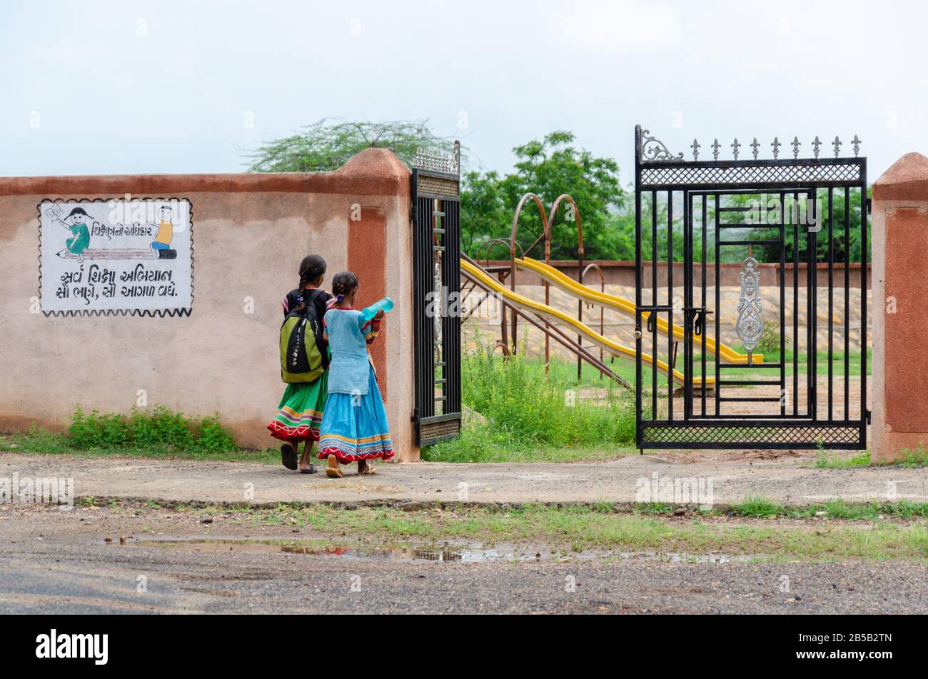 Due bambine che camminano con la borsa di scuola, entrando nel cancello della scuola contrassegnato da Sarva Shiksha Abhiyan logo a Gandhi Nu Gam, Kutch, Gujarat, India Foto Stock