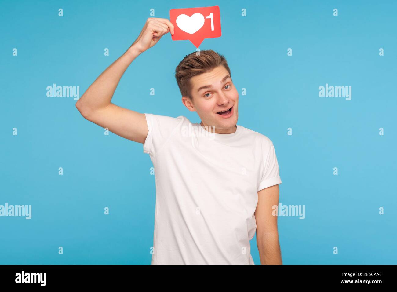 Uomo sorridente spensierato in t-shirt bianca tenendo il tasto social media sopra la testa, mostrando cuore come icona, dando consigli per il supporto dei contenuti, fo Foto Stock
