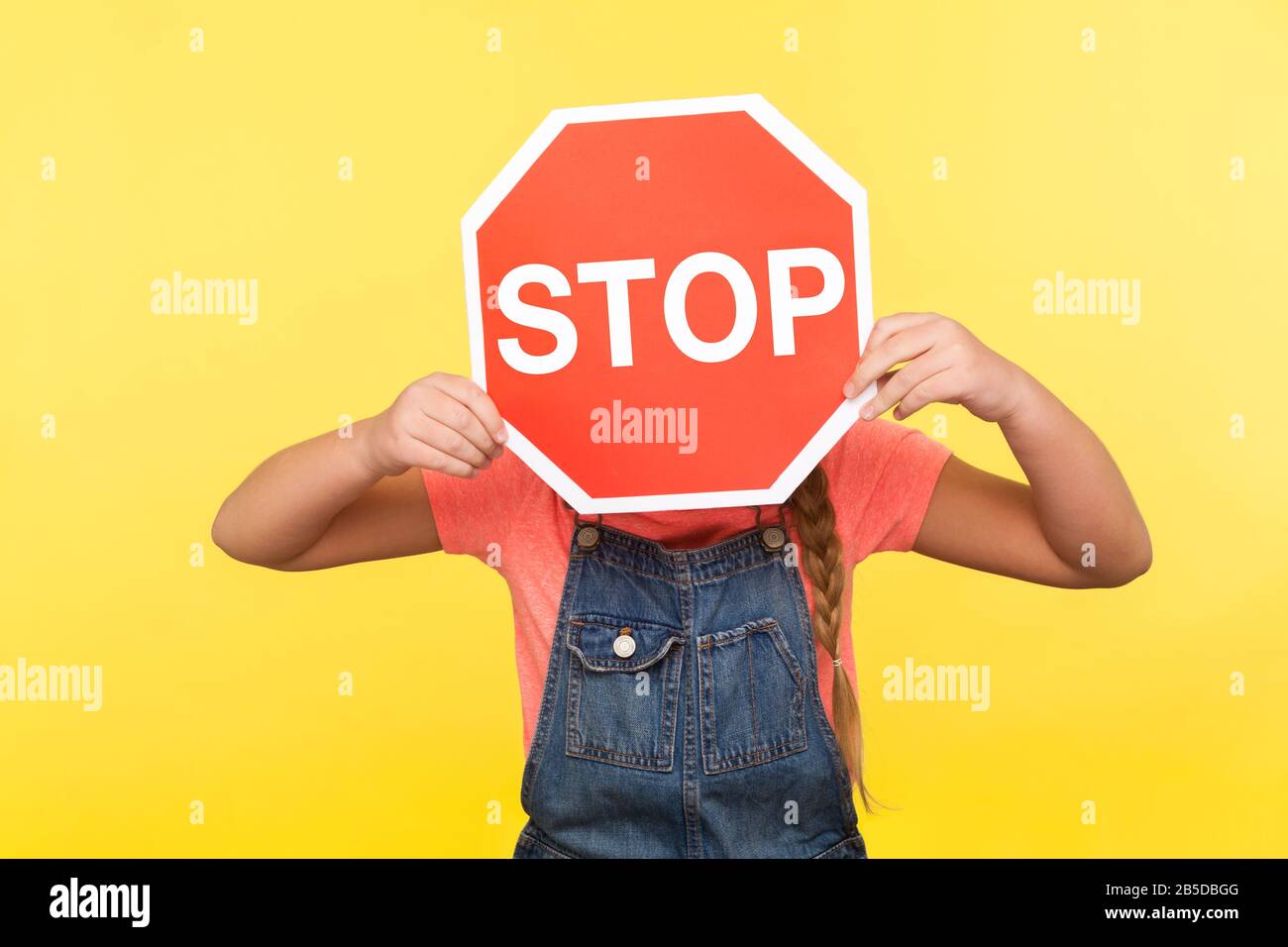 Modo proibito. Bambina in tute in denim che copre il viso con il simbolo di arresto ottagonale, tenendo il segnale del traffico rosso, attenzione al passaggio sicuro della strada. IND Foto Stock