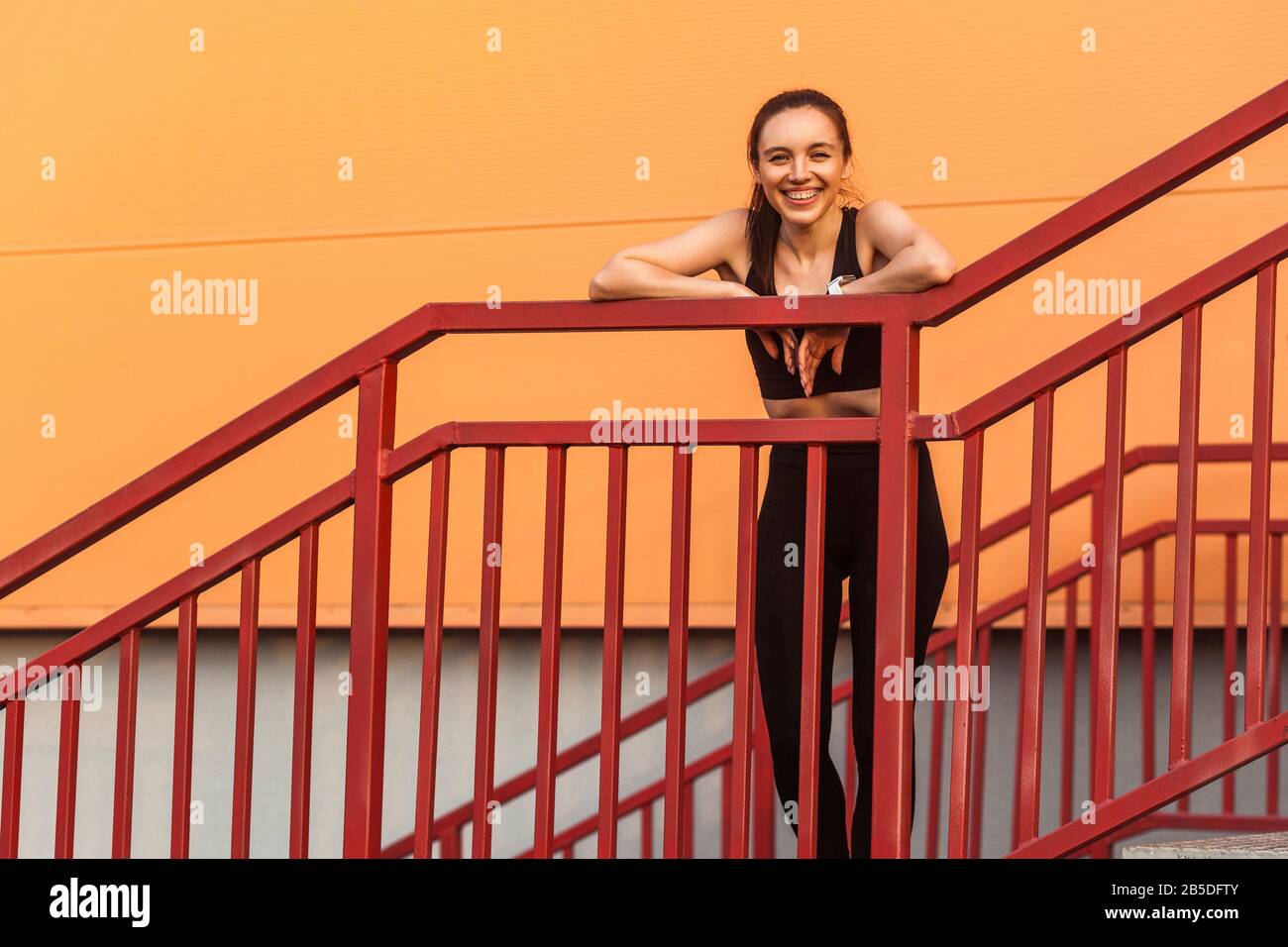 Indossa una ragazza slim-positive in pantaloni sportivi e la parte superiore poggiata sulle scale dopo il jogging e guardando la fotocamera con sorriso, facendo una pausa durante l'allenamento di corsa o Foto Stock