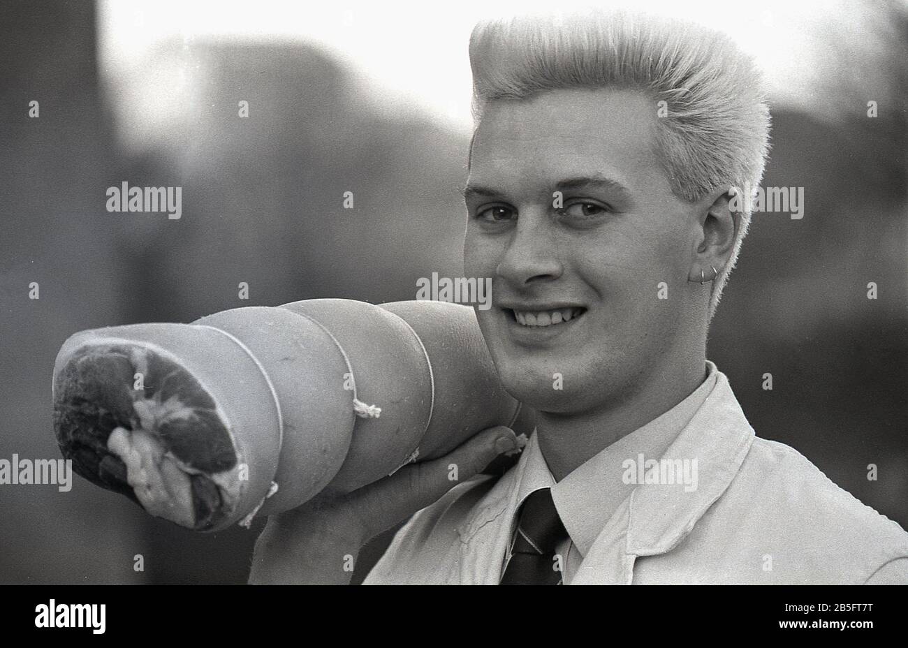 Anni '1980, storico, un giovane macellaio maschio con taglio di capelli spiky e orecchini che porta una gamba di maiale sulla spalla, Yorkshire, Inghilterra. Foto Stock