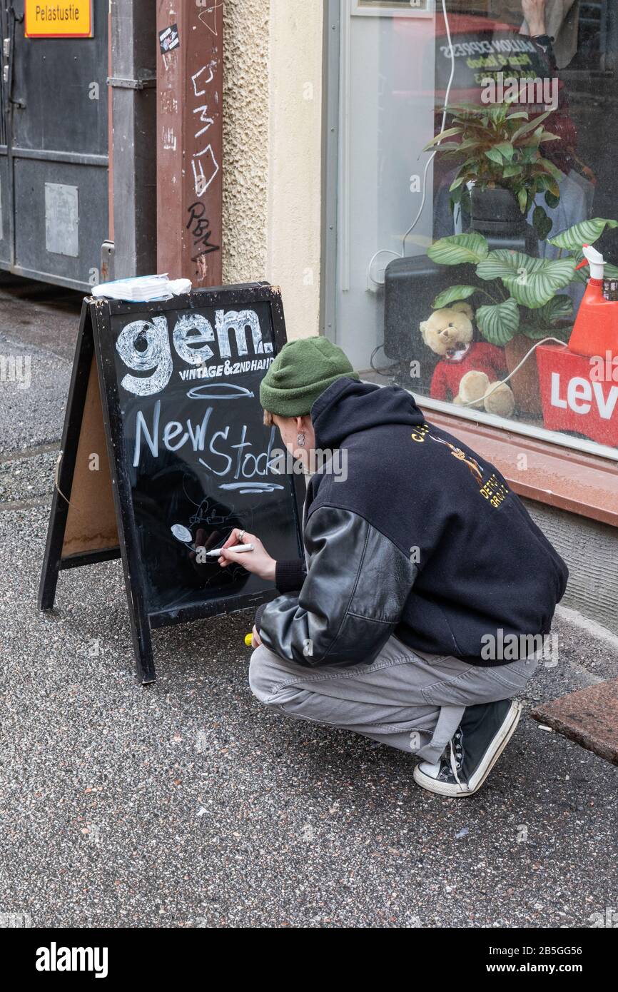 Giovane uomo che scrive su una lavagna marciapiede di fronte a Gem vintage e negozio di seconda mano nel distretto di Kallio di Kallio, Finlandia Foto Stock
