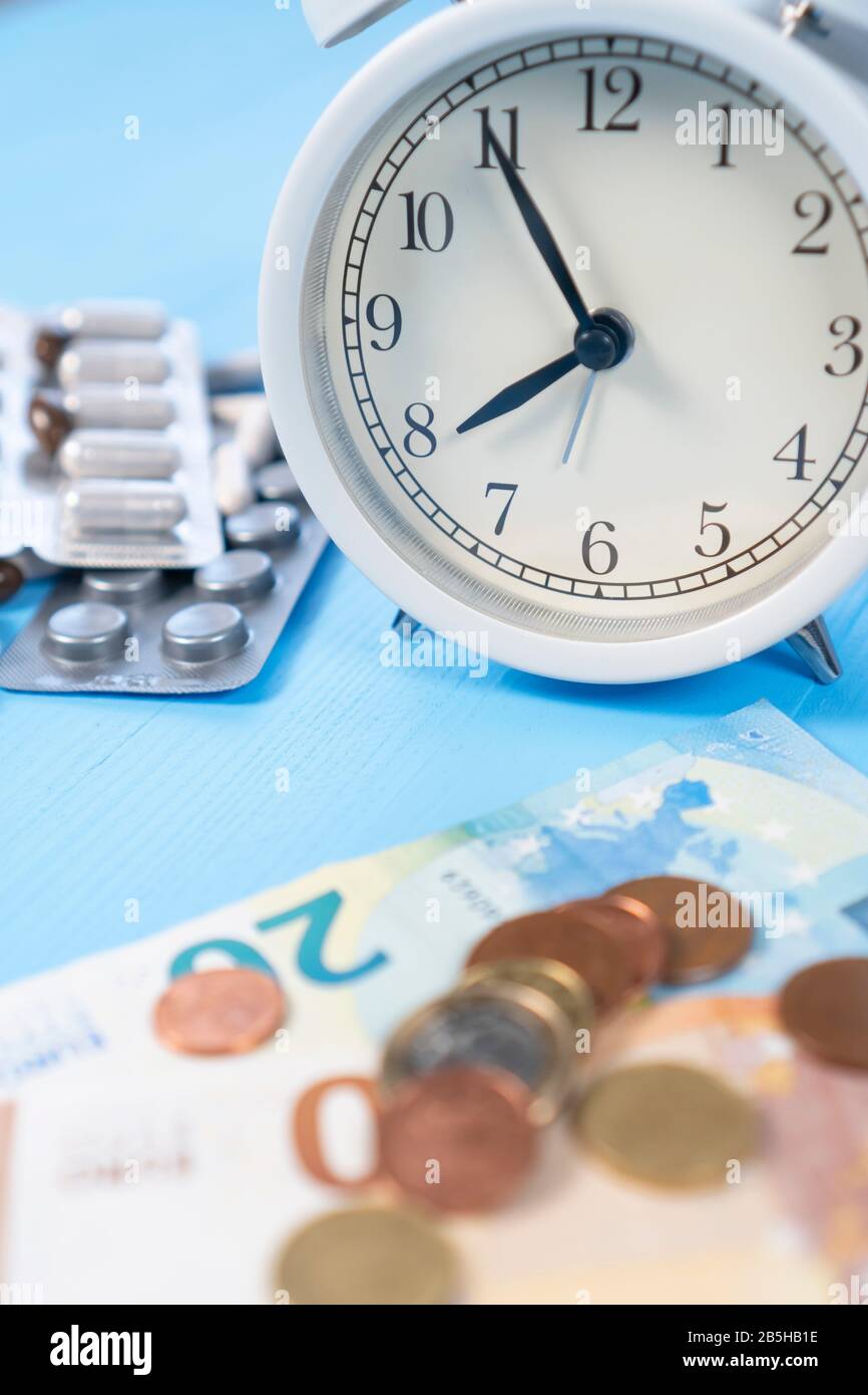 Banconote e monete in euro con un mazzo di pillole mediche con sveglia bianca su sfondo di legno azzurro. Vista verticale. Concetto di problemi Foto Stock