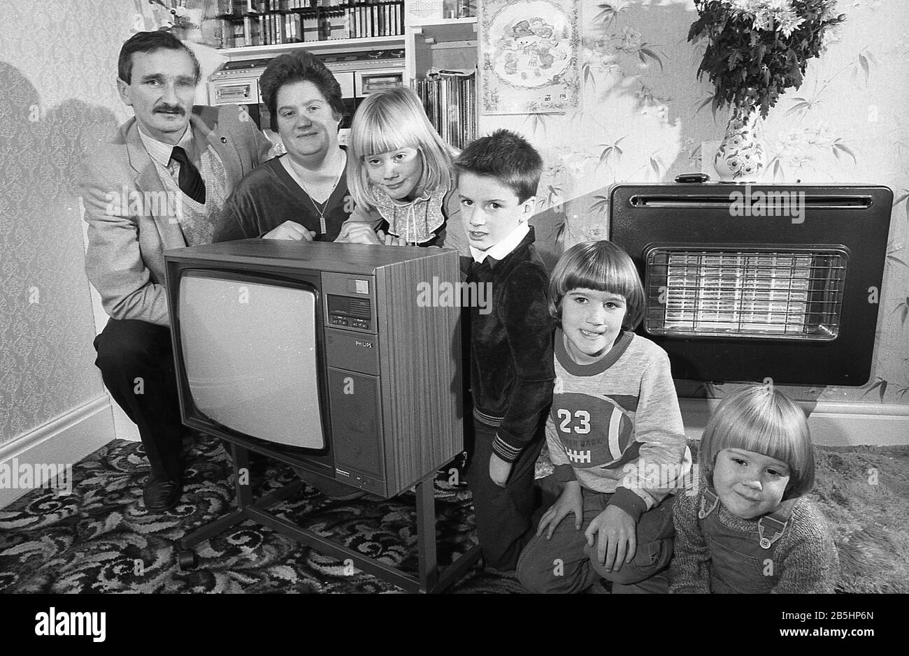 Anni '1980, storico, all'interno di una sala d'ingresso, una famiglia, una madre, un padre e i loro quattro figli piccoli, si schierano per una foto, accovacciandosi accanto alla loro nuova televisione seduto su un piedistallo di metallo con ruote, Inghilterra, Regno Unito. Un fuoco da bar elettrico montato a parete dell'epoca visto nella foto. Foto Stock