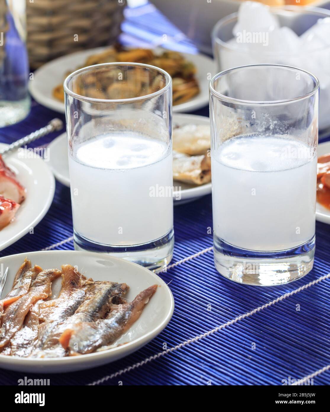 Ouzo, raki alcol con cubetti di ghiaccio accanto a meze di pesce su piatti bianchi. Due bicchieri e vari molluschi sani, vista ravvicinata, verticale. Foto Stock