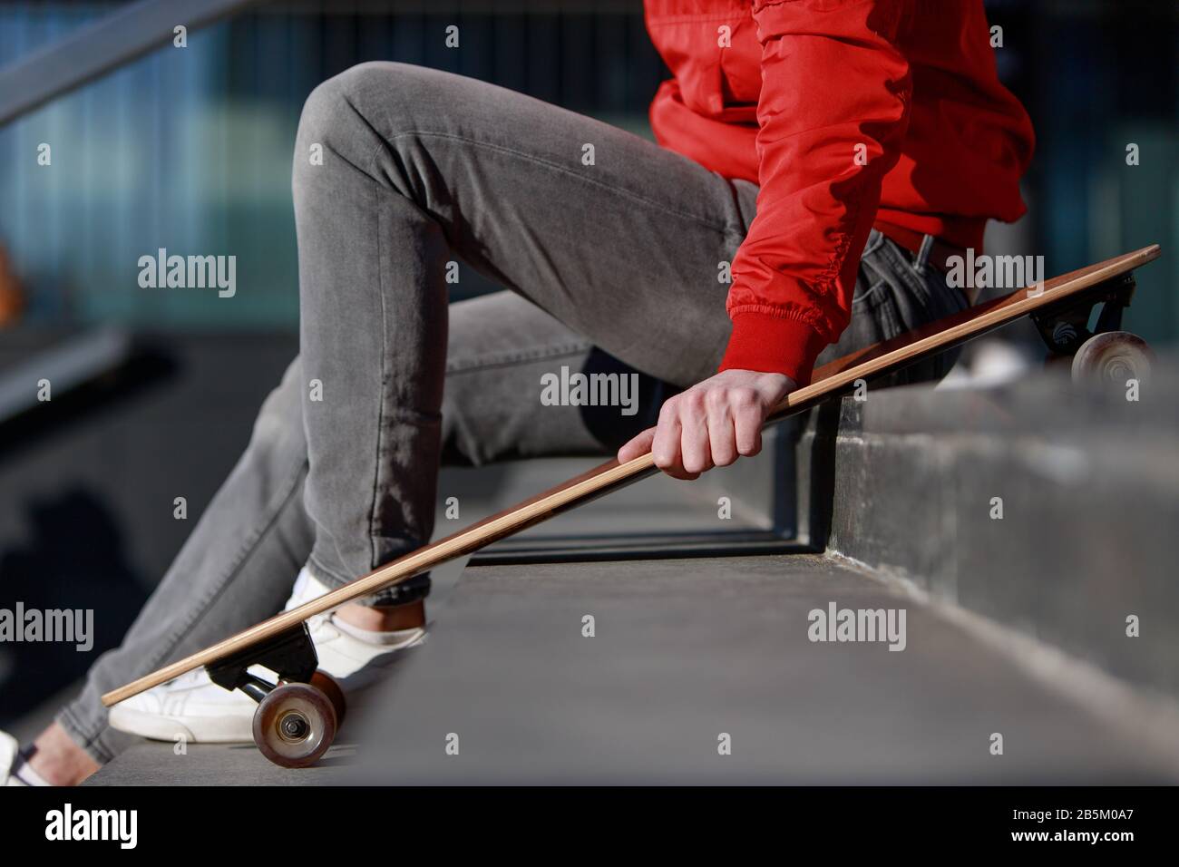 Elegante longboarder uomo in abiti casual appoggiato sui gradini, seduto con longboard/skateboard all'aperto, vista laterale, immagine ritagliata. Urbano, subcultur Foto Stock