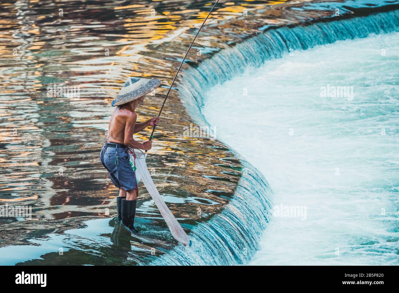 Pescatore con un tradizionale cappello cinese triangolare in piedi nelle acque del fiume Tuo che scorre attraverso il centro della città antica Fenghuang Città Vecchia Foto Stock