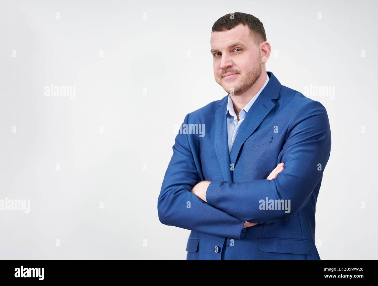 Giovane uomo d'affari di successo con le braccia incrociate che indossano vestito blu con un sorriso genuino grazioso che posano contro lo sfondo grigio con lo spazio della copia Foto Stock